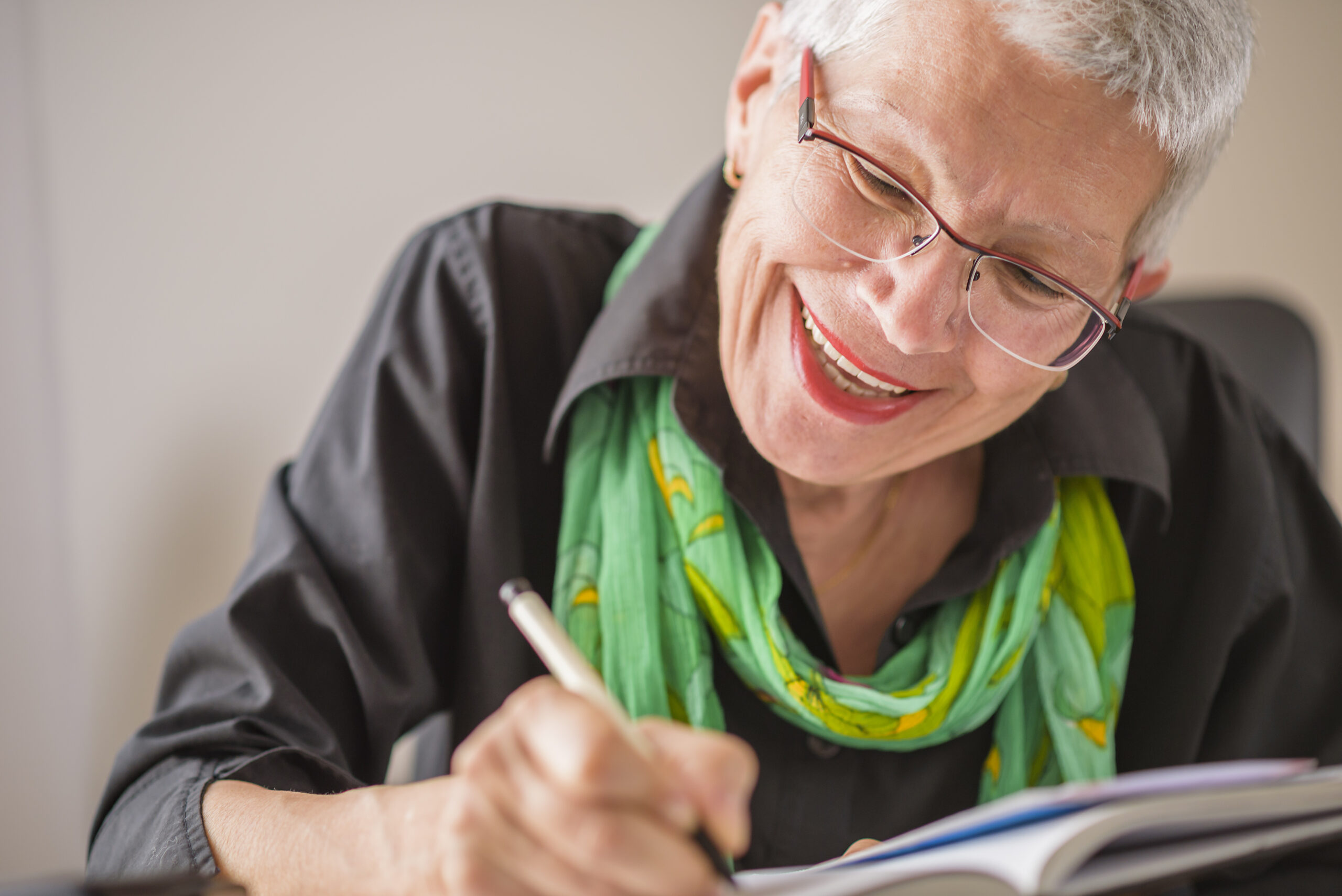 Mature woman with glasses and white hair journaling happily.